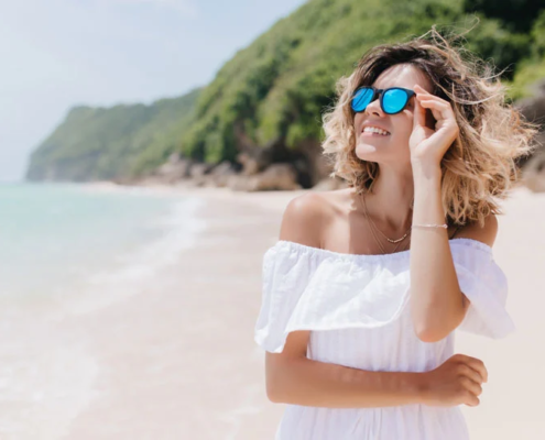 Haarpflege nach dem Strandurlaub - junge Frau mit Sonnenbrille am Strand.